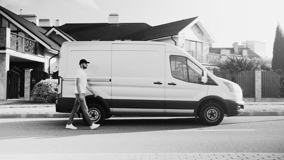 A man wearing a black cap, white t-shirt, and jeans is outdoors, loading three stacked cardboard boxes into the side of a white cargo van parked on a residential street in Church End. The van, associa