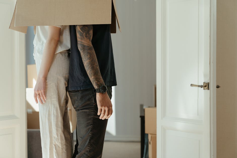 A man with a tattooed arm wearing a black T-shirt and black pants is seen during a house move, carrying a large cardboard box into a room with white paneled doors. An individual with lighter clothing
