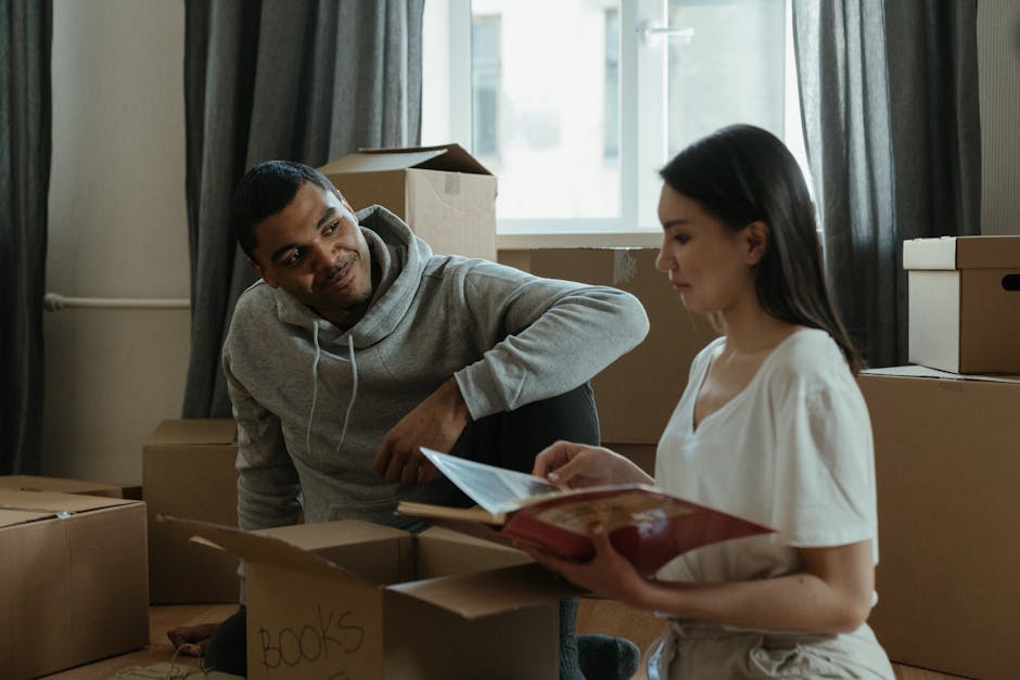 A man with a tattooed arm wearing a black T-shirt and black pants is seen during a house move, carrying a large cardboard box into a room with white paneled doors. An individual with lighter clothing