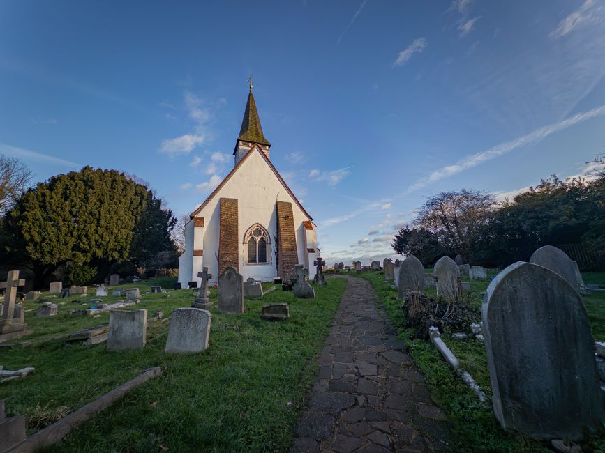 A view of a small, white church with a tall, pointed steeple set within a graveyard, featuring numerous weathered headstones arranged on either side of a narrow, uneven stone pathway. The sky above is