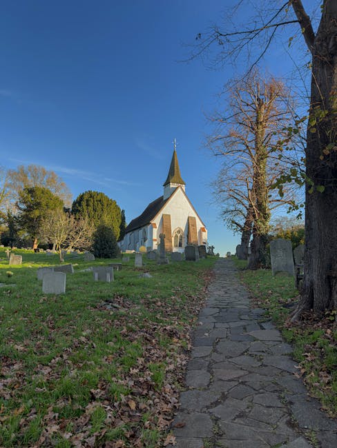 A narrow stone pathway leading uphill through a churchyard, with uneven paving stones on a slightly inclined terrain surrounded by grass, fallen leaves, and mature deciduous trees with bare branches, indicating late autumn or early winter. To the left, there are several tombstones of varying sizes and shapes, positioned at irregular intervals, some partially obscured by grass. The pathway ascends towards a small white church with a dark, steeply pitched roof, a tall, pointed steeple topped with a metal cross, and narrow arched windows. The scene is illuminated by daylight under a clear blue sky, and the background features additional trees with dense green foliage and a few distant monuments. This setting illustrates a typical countryside churchyard suitable for house removals and furniture transport, with [COMPANY_NAME] providing relocation services, involving careful loading and moving logistics around such historic locations.