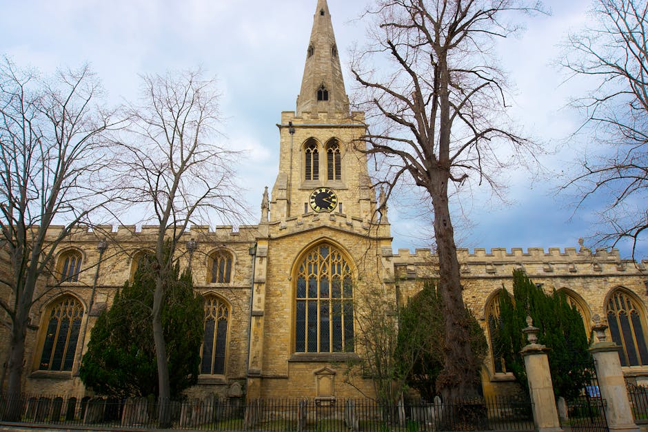 An exterior view of a historic stone church with tall, arched stained glass windows and a prominent clock on its tower, set against a partly cloudy sky. Surrounding the church are leafless trees and evergreen shrubs, with a low metal fence at the front. The church’s architecture features crenellated battlements along the roofline and a steeply pointed spire, indicating a Gothic style. The scene captures the building during daytime, with natural light illuminating the stonework. This setting relates to home relocation and moving services provided by Man with Van Church End, especially in the context of relocating items from or to such institutional buildings, with the loading area near the entrance visible for furniture transport and packing processes.