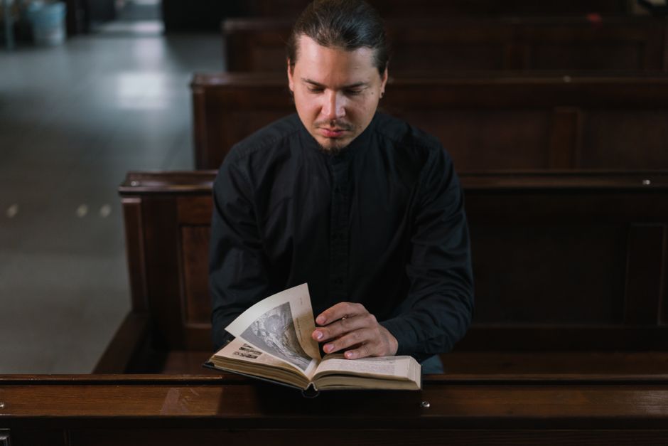 Inside a church with high ceilings and wooden pews, a man dressed as a priest in a black clerical shirt with a white collar is seated on a pew, facing a young woman with long brown hair wearing a beig