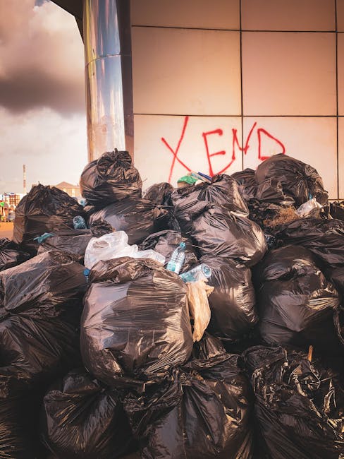 A large pile of black rubbish bags filled with waste, situated on a pavement against a tiled wall with red graffiti spelling 'XEND'. Several plastic bottles are visible within the bags, some partially protruding. The bags vary in size and appear to be tightly sealed. Behind the pile, part of a modern, reflective building surface is visible, with a cloudy sky overhead. The scene depicts waste disposal or clearance related to house removals or bulky waste removal services offered by Man with Van Church End, with the focus on waste collection and transport logistics for home relocation projects.