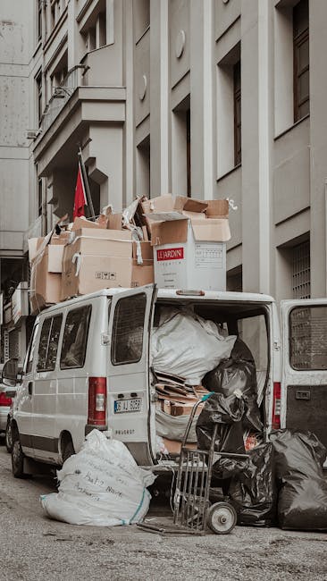 The rear of a white van parked on a city street next to a multi-storey building features an open rear door revealing a loading area filled with large black and white rubbish bags, flattened cardboard boxes, and wooden pallets. Several cardboard boxes, some labeled and others unmarked, are stacked on top of the van's roof, with additional boxes and packaging materials visible inside through the open tailgate. A small platform trolley is positioned on the pavement beside the van, loaded with black rubbish bags. The scene appears to be part of a home relocation or furniture transport process, with the van used for moving bulky waste or household items. Man with Van Church End specializes in removals and waste collection, supporting efficient packing and moving logistics, as seen in the organized loading environment and the equipment present for handling large items and waste disposal.