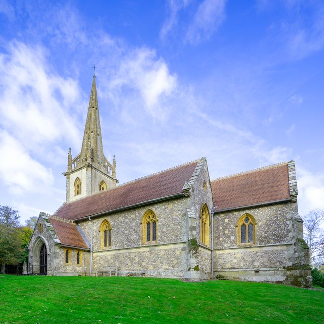 Photograph of a historic stone church with a tall, pointed spire and arched windows, situated on a grassy area with a gentle slope. The church has a red-tiled roof, and its exterior features weathered