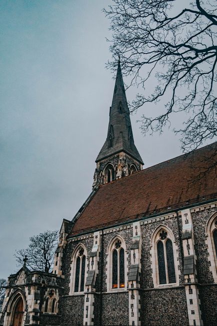 A view of a historic stone church with a tall, pointed spire under a cloudy sky, featuring arched stained glass windows and decorative stonework. Bare tree branches extend into the frame from the right side. The church is situated in an outdoor setting, with no visible vehicles or moving equipment. The scene captures the exterior of the building, emphasizing its architecture and surroundings, relevant to local house and church removal services by Man with Van Church End, illustrating the type of property involved in relocation or moving processes.