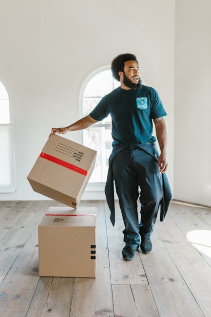 A man with light skin, wearing a white polo shirt, sunglasses, and a cap, is seen loading cardboard moving boxes into the back of a white moving van during a home relocation. The van's open rear door