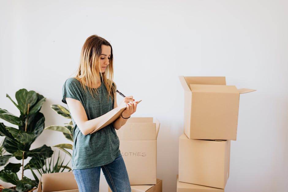 A young woman with long blonde hair, dressed in a casual dark green t-shirt and blue jeans, is standing indoors next to several stacked cardboard boxes, some open and others sealed with tape. She is holding a clipboard and pen, appearing to take notes or check inventory as part of a home relocation or packing process. In the background, there is a large green potted plant with broad leaves, positioned against a plain white wall. The scene suggests an organized packing or moving preparation, with visible materials such as cardboard boxes and packing tape, indicative of a professional removals or furniture transport service, like those offered by Man with Van Church End, involved in home moving or house removal logistics.