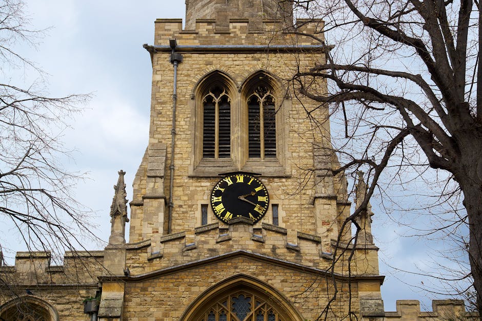The image shows the exterior of a historic stone church tower set against a cloudy sky, with leafless tree branches partially framing the sides. The tower features two tall, narrow arched windows with wooden louvered shutters near the top, and below them is a round black clock face with gold Roman numerals and hands displaying the time. A stone statue is situated on the left side of the tower's ledge. The structure appears to be part of a larger church building with gothic architectural elements, and the scene is captured during daytime with soft natural light illuminating the stonework. The context of the image relates to traditional buildings, which could be associated with a house removals or moving service when considering the seamless integration of historical architecture typical in London or UK settings, such as those serviced by Man with Van Church End for home relocation and furniture transport.