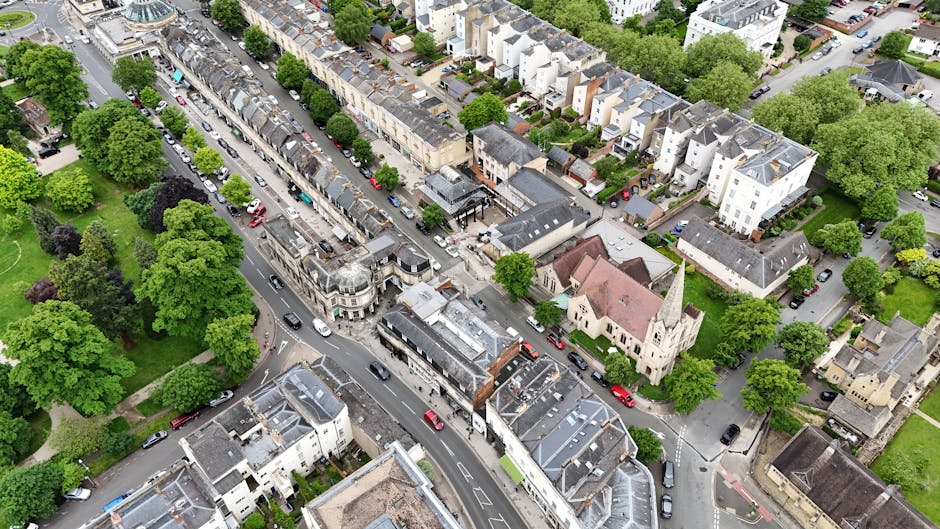 An aerial view of a residential street in Church End, featuring a mix of terraced and semi-detached houses with grey and white facades, surrounded by lush green trees and small gardens. A church with a tall spire is visible among the buildings. The street includes parked cars along both sides, with some vehicles on the pavement and others on the road. In the foreground, a large white van with open rear doors is parked partially on the sidewalk, indicating a loading process for a home relocation or furniture transport. The van is equipped with protective blankets and straps for moving furniture. Nearby, stacks of cardboard boxes, packing materials, and wrapped furniture are visible, suggesting an ongoing packing and loading operation by a professional removals company, such as Man with Van Church End, preparing for a same-day emergency move. The lighting is natural, indicating daytime in clear weather.