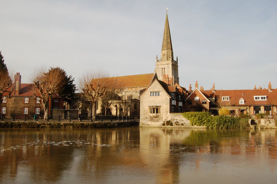 The image shows the exterior of a historic stone church tower set against a cloudy sky, with leafless tree branches partially framing the sides. The tower features two tall, narrow arched windows with wooden louvered shutters near the top, and below them is a round black clock face with gold Roman numerals and hands displaying the time. A stone statue is situated on the left side of the tower's ledge. The structure appears to be part of a larger church building with gothic architectural elements, and the scene is captured during daytime with soft natural light illuminating the stonework. The context of the image relates to traditional buildings, which could be associated with a house removals or moving service when considering the seamless integration of historical architecture typical in London or UK settings, such as those serviced by Man with Van Church End for home relocation and furniture transport.