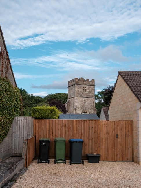 A wooden boundary fence surrounds a small outdoor area with four waste bins in front of it—black, green, blue, and a smaller black bin—positioned on a gravel surface. To the left, part of a brick wall with greenery is visible, while a narrow surface with stacked paving stones runs along the left edge. Behind the fence, a historic stone tower with battlements and small windows is seen, set against a partly cloudy sky with patches of blue and white clouds. On the right, the side of a residential building with a tiled roof is partially visible. In this scene, Man with Van Church End is preparing for a home relocation involving packing and loading furniture and boxes, with the outdoor area ready for vehicle access and the collection of household items for a furniture transport or removal service.