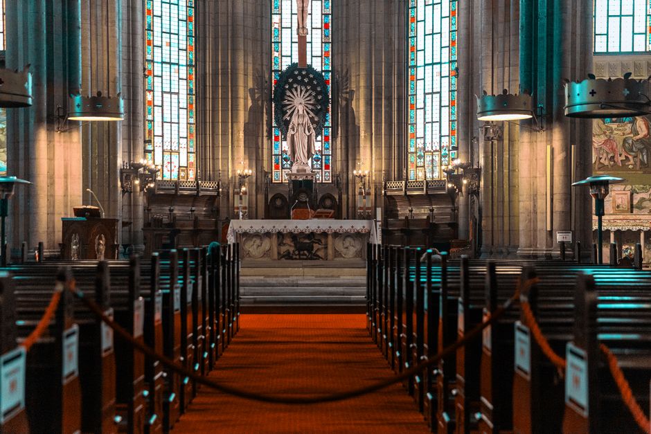 Inside a church with an ornate altar centrally positioned, featuring a large religious statue, surrounded by tall stained glass windows with vibrant red, blue, and yellow hues. The interior is illuminated by natural light filtering through the windows and electric chandeliers hanging from the high ceiling. The nave is lined with dark wooden pews, separated by orange-and-brown carpeted aisles sectioned off with orange ropes attached to black posts, indicating a restricted area. Near the front, on the left side, a cart with cardboard boxes and packing materials is partially visible, suggesting preparations for a move or recent relocation. Behind the pews, a small area contains additional boxes, plastic wrapping, and a trolley, indicating ongoing packing or furniture transport processes related to house removal or moving logistics. Overall, the scene captures a moment of organization within a historic church, with attention to safe removal practices and packing materials, consistent with house removals and relocation services offered by Man with Van Church End.